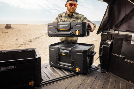 StackTech Campingbox von ToughBuilt im Autokofferraum am Strand, umgeben von Sand und Wellen, ideal für Camping und Outdoor.