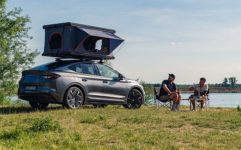 Ein Skoda Enyaq steht in einer Seen Landschaft, ausgestattet mit einem Campnest 120 Dachzelt von mycamperbox. Das Dachzelt in modernen, minimalistischen Farben ist aufgeklappt, und warme Sonnenstrahlen aus dem Zelt. Der SUV ist umgeben von einem Strand und großen Bäumen, während im Vordergrund Camping-Equipment wie ein kleiner Tisch und Stühle zu sehen sind. Der Gesamteindruck vermittelt eine Kombination aus Funktionalität und Komfort für das Beachcamping.